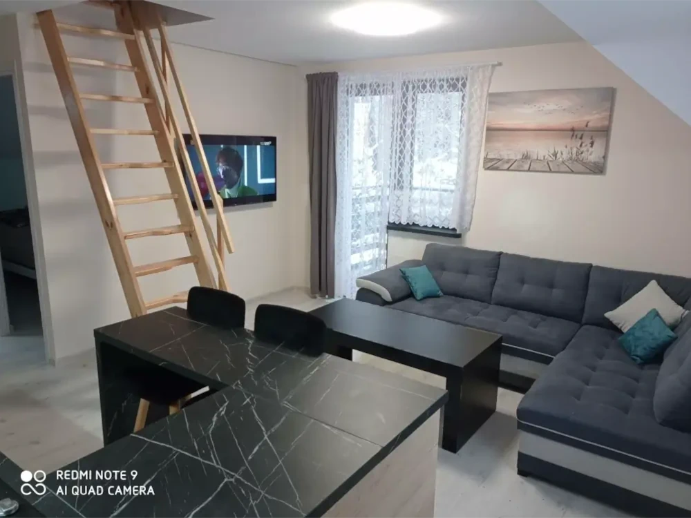 Salon z jasnym oświetleniem, telewizorem, wyjściem na balkon i schodami prowadzącymi na poddasze